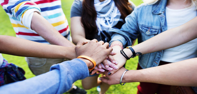 Group of friends placing hands together in a circle on a grassy field to symbolize teamwork and unity.