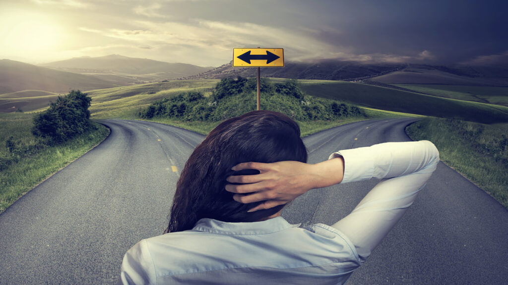 Person seen from behind at a fork in a rural road, hands on head, contemplating which direction to take.
