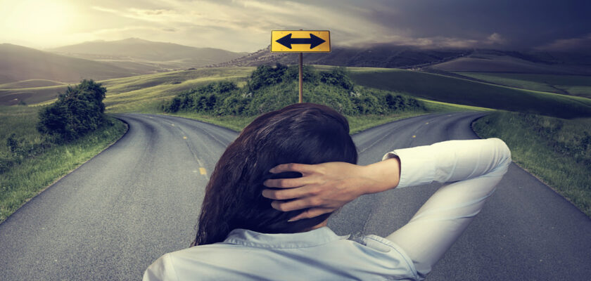Person seen from behind at a fork in a rural road, hands on head, contemplating which direction to take.