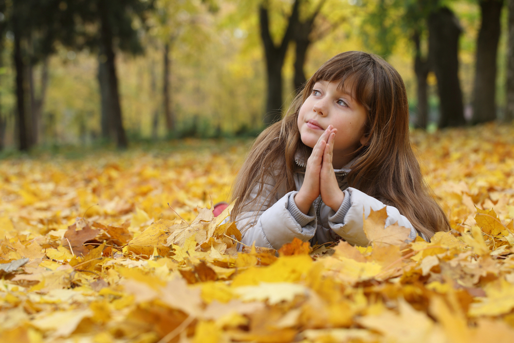 Young girl lies in a bed of yellow autumn leaves, hands pressed together in a prayer-like pose, looking upward.