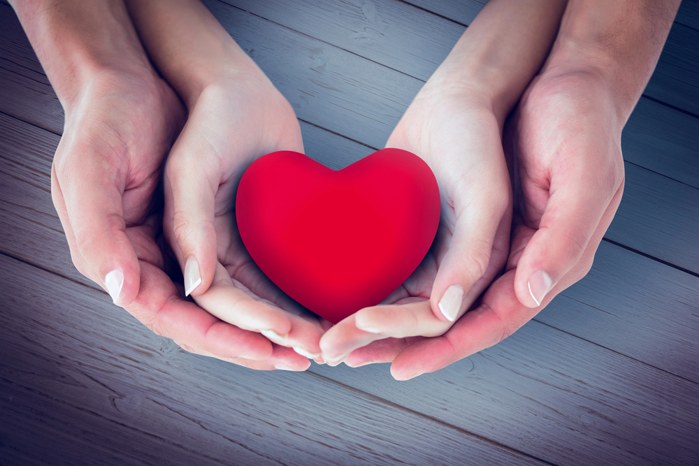 Two pairs of hands gently cupping a red heart, symbolizing care and compassion on a wooden surface.
